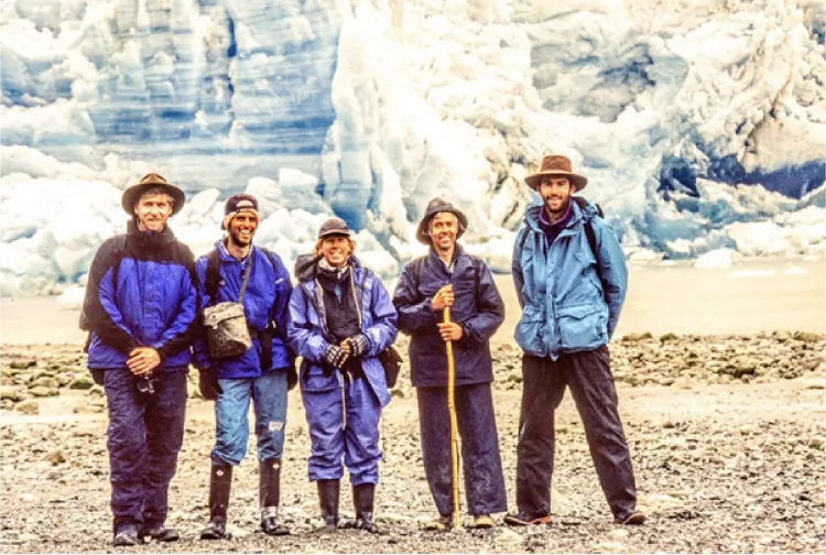 five people in blue rain gear smiling in front of a blue-white glacier