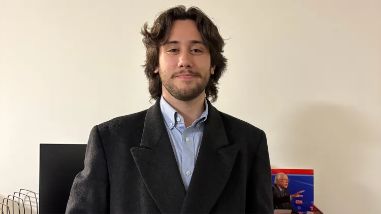 Young man with brown hair and beard, wearing dark suit and light blue shirt, standing in front of large computer monitor and laptop showing Senator Bernie Sanders speaking on CNN