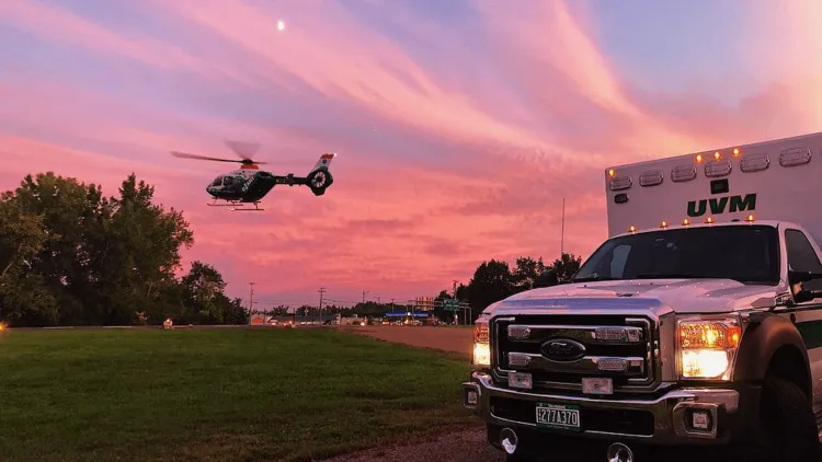 A UVM ambulance parked near a hovering helicopter against a colorful sky