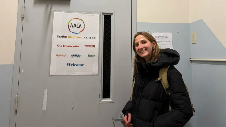 Young woman with long dark blonde hair and black winter coat standing in front of gray door with a sign that says "AALV Inc." and has the word "Welcome" written in nine languages