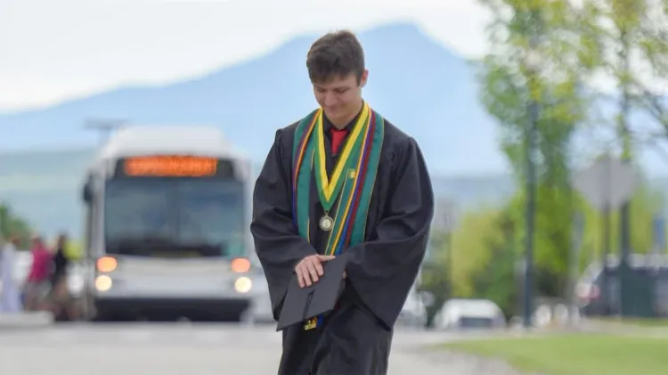 Graduate in a black gown and colorful honor cords holding a cap while walking outdoors