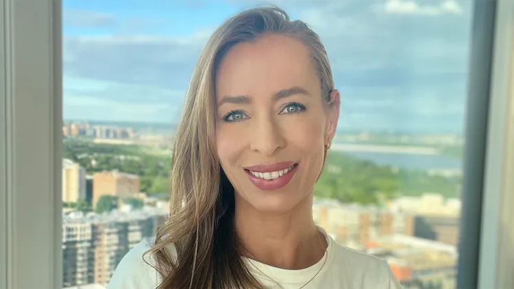 Person smiling in front of a window with a cityscape in the background.