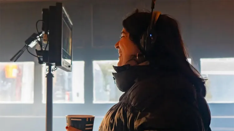 Person smiling while wearing headphones and standing beside a monitor on a film set.