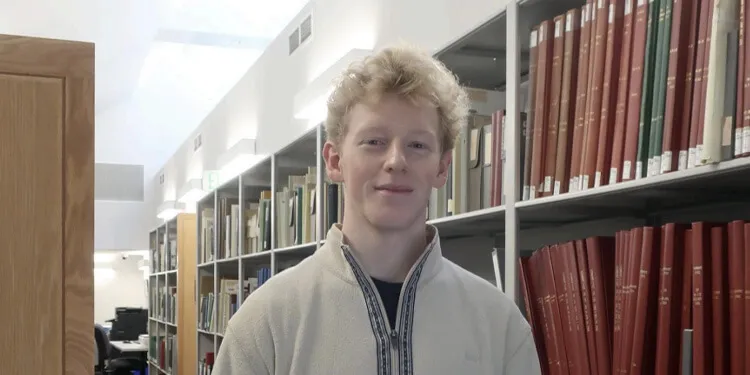 A person standing in a library, smiling while surrounded by bookshelves.