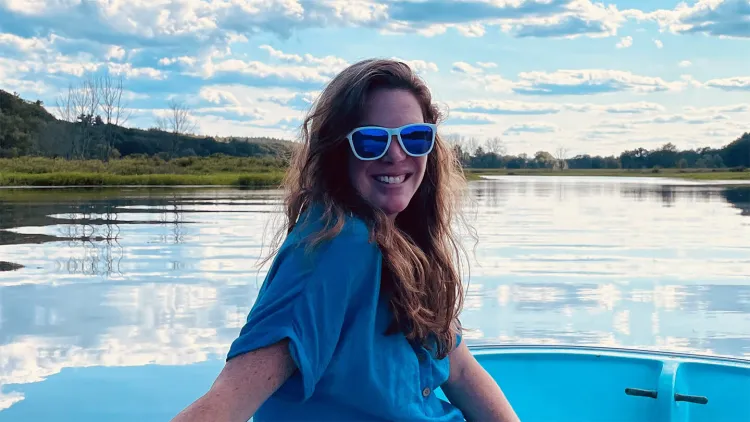 Person wearing sunglasses smiling while sitting on a boat on a calm lake.