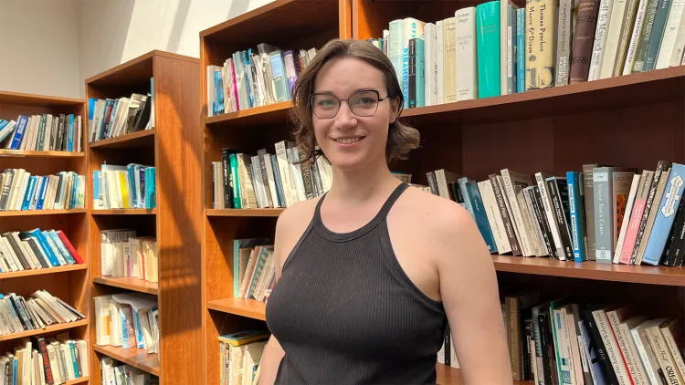 Person smiling while standing in a sunlit library, surrounded by bookshelves.