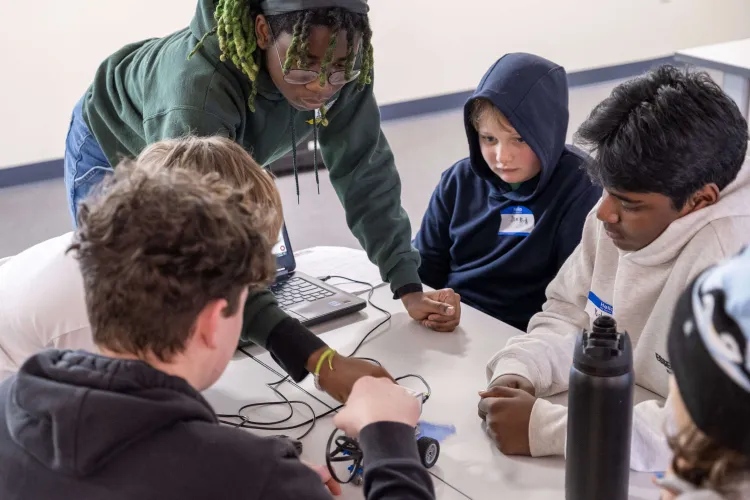 An older student standing over a table of younger students and demonstrating with their hands with a small machine hooked up to a laptop.