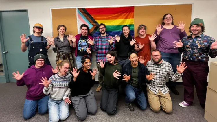 Fourteen individuals, a mix of UVM professors, graduate students, and undergraduates, posing with their hands out for a photo in front of a pride flag.