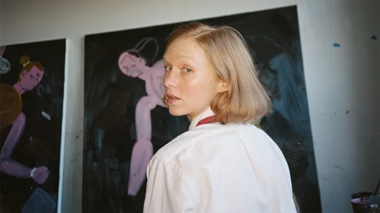 A person stands in an art studio with large paintings displayed on the wall behind them.