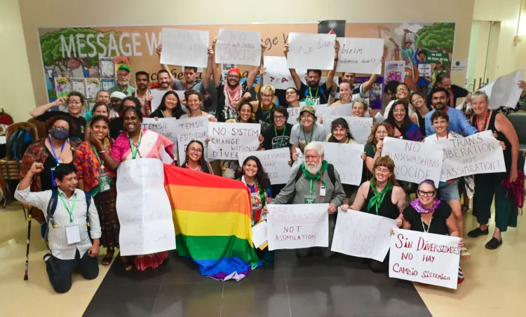 A group gathers holding signs and a rainbow flag for a diversity march at the Nyéléni Global Forum. Signs say, “no system change without diversity” “no pink washing genocide” and “trans liberation not assimilation”.