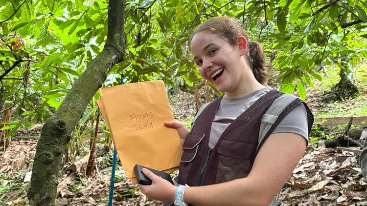A smiling person standing in a leafy forest, wearing a vest and backpack, holds up a manila envelope labeled “Frogs data” beside a tree.