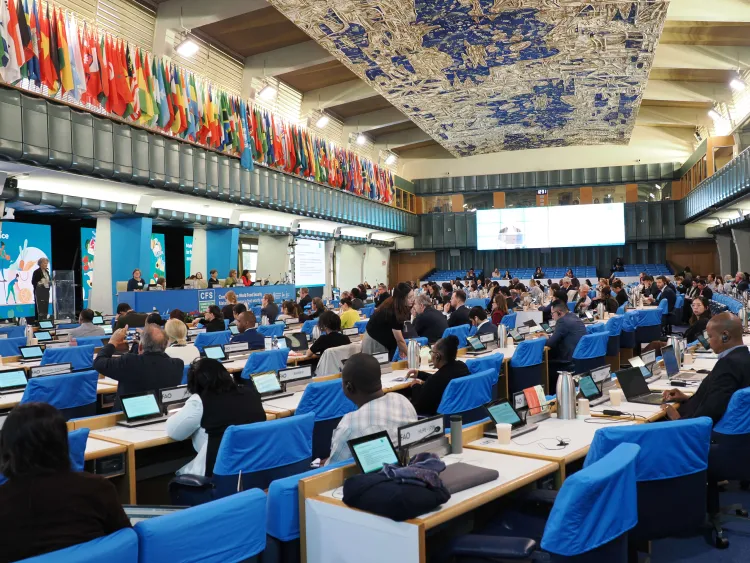 Committee room with blue chairs, many occupied, with flags adorning the walls