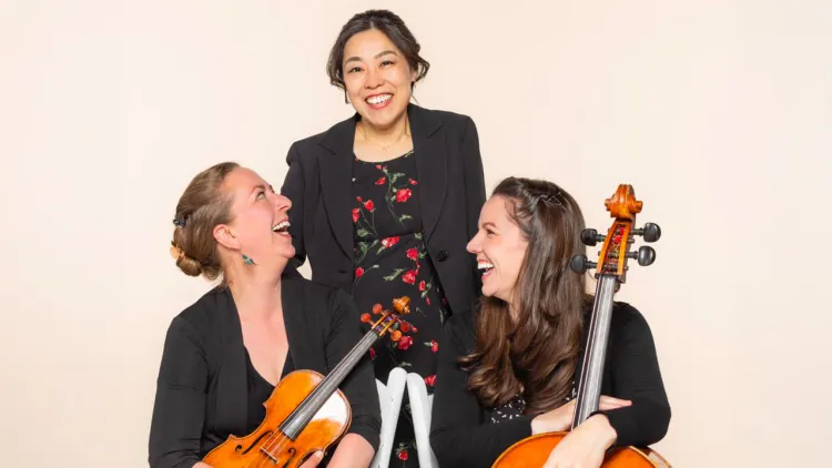 Two people are seated holding a violin and a cello, while one person stands between them.