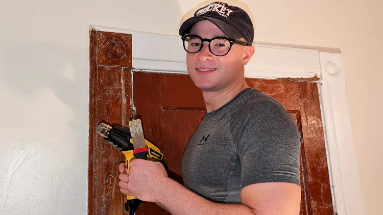 Person in glasses and a cap holding a heat gun by a wooden doorframe.