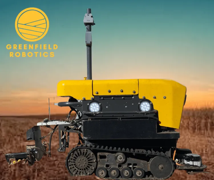 A yellow robot with rubber tracks against a blue and orange sunset sky. In the top left corner is a yellow round logo that says greenfield robotics. 