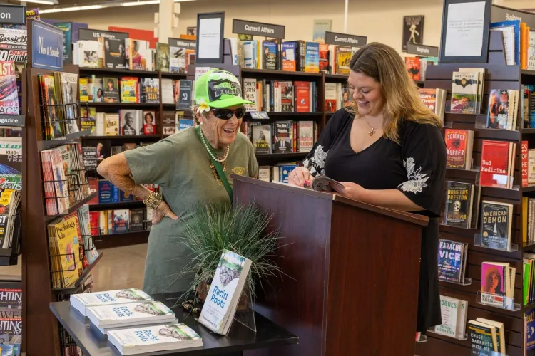 Woman at wooden podium smiles next to woman in green shirt in hat inside a bookstore.
