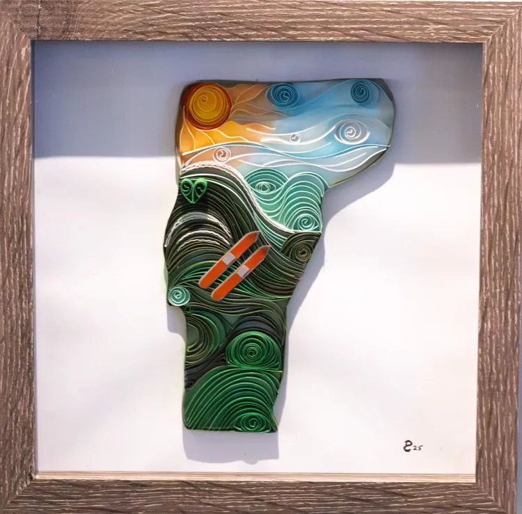Framed quilling design of the state of Vermont, made with colored paper. The shape of the state of Vermont has an abstracted landscape inside it, made of rolled paper. There is a yellow and orange sun in the top left corner, with blue swirls flaring out from the sun and toward the right. Below this there are rolling green hills with a pair of red skis on in the center of them.