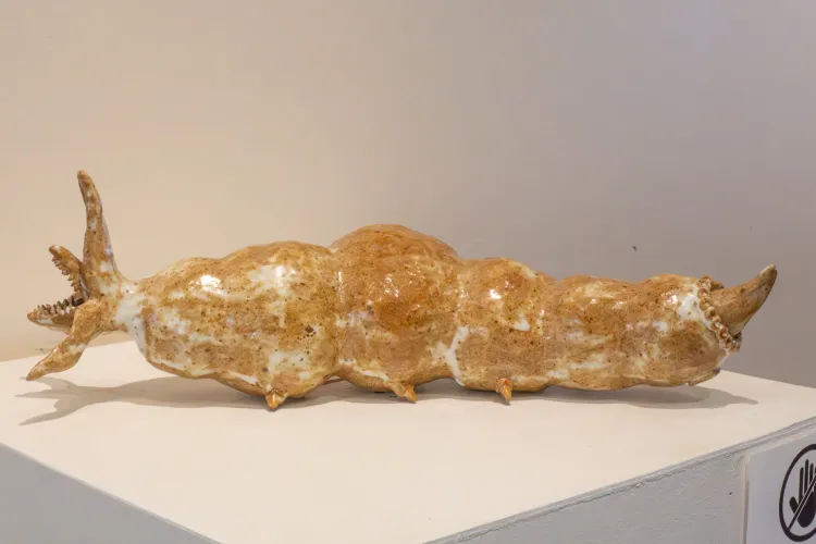 Glazed ceramic sculpture of a worm-like creature. It is a tan and cream speckled creature with a bubbly worm-like body shape. It has several small stubs for legs and a pointed end like a tail. It's head is a three pronged opening with a mouth protruding with many small sharp teeth. 