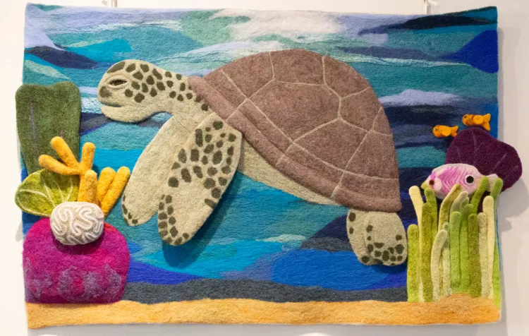 A felted oceanic scene of a turtle swimming among coral and other sea creatures. The turtle swims through the center of the wall hanging, framed by purple and green coral. Two goldfish and a purple fish swim to the right of the turtle. The background is made of a variety blue, teal and white felt to represent the ocean. There is a strip of tan felt along the bottom of the piece to represent the sand of the ocean floor. 