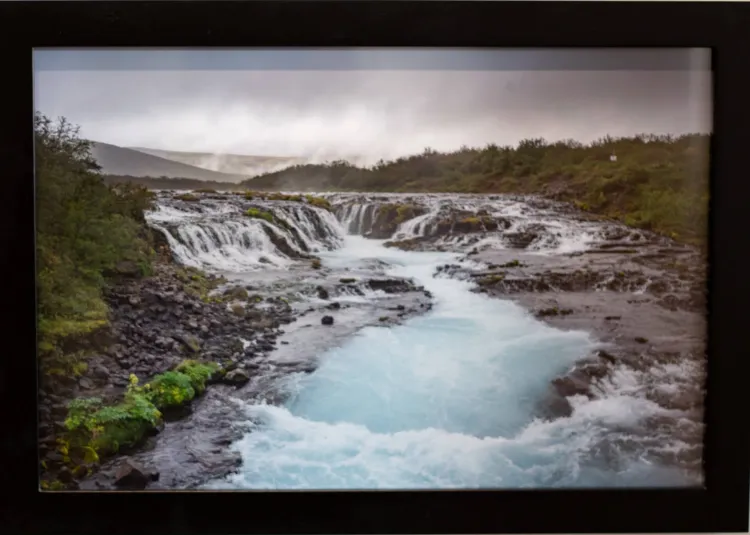 Framed landscape photograph of the Brúarfross Waterfall in Iceland. It is a cloudy scene over the waterfall, which is centered in the photograph with rushing light blue water running down a rocky path. On either side of the waterfall there are mossy areas with green trees. The sky is gray like the rocks with light coming in near the horizon.