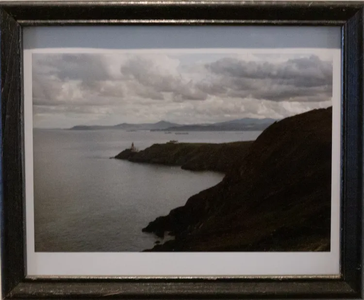 Framed photograph of an oceanic landscape, featuring a lighthouse at the end of a grassy hillside. In the foreground, hills on the right decline to meet a teal ocean on the left.  Part of the hill continues to the center of the photograph, with a lighthouse on the end of the plateau. It's a cloudy scene and there are more hills in the background.