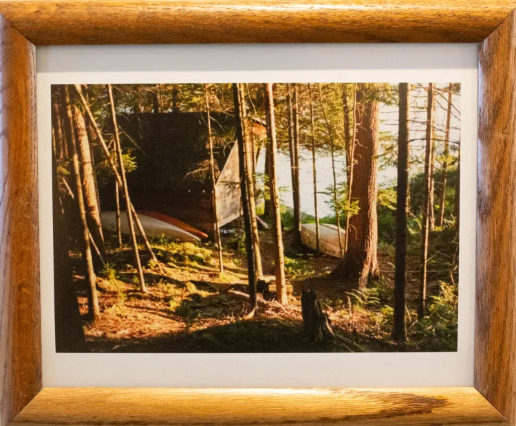 Framed photograph of a cabin beside water in a forest, with boats resting beside the cabin. There are trees in the foreground, with light coming in from beyond the image to the right. The back of a cabin rests on the left of the image, with two kayaks just before it and a small dingy to the right of it. The blue water of a lake begins to the right of the lake, which can be seen through the trees. 