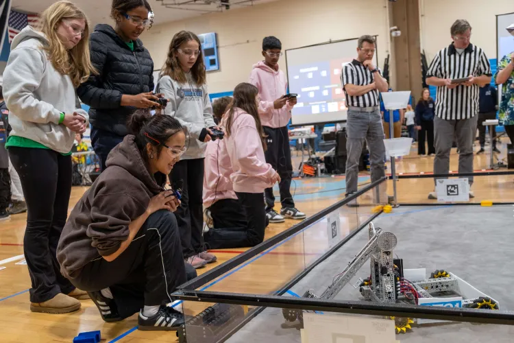 Seven young people in clear safety glasses and two adults in black and white striped referee shirts stand around an enclosure, looking at a robot