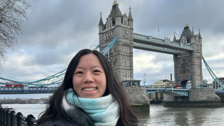 Person smiling in front of Tower Bridge in London on a cloudy day.