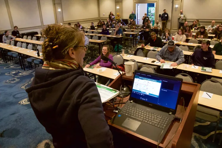 An image of a person standing at a podium with a room full of people sitting at long tables smiling back. 