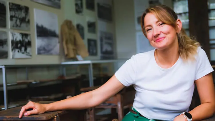 Person seated indoors beside a wooden display case with framed photographs on the wall behind them.