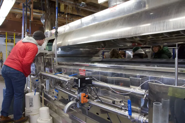 A person in a red coat and hat leans over a large metal container, an evaporator.