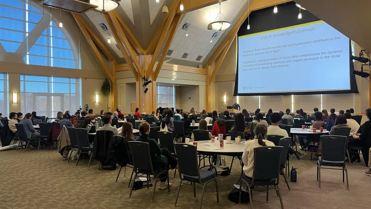 A large ballroom with many individuals giving their attention to a presenter and their slides at the front of the room.