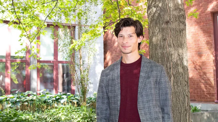 Person standing outdoors near a tree, smiling, with greenery and a brick building in the background.