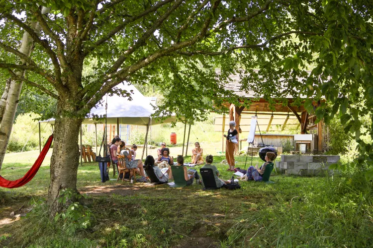 People sit in a circle under trees with tent in the background
