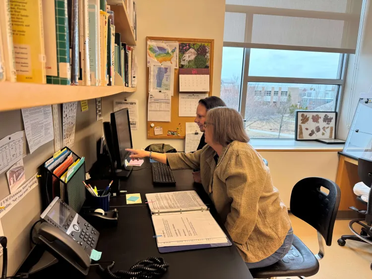 Two people sit at a desk with books on the wall and a computer screen and telephone in front of them
