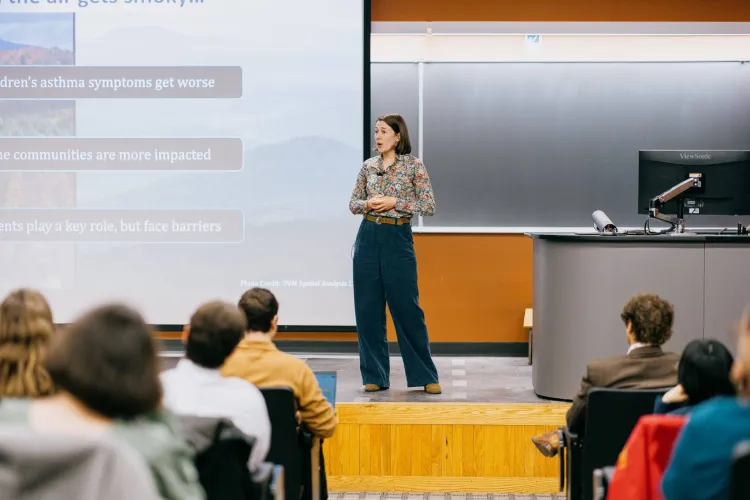 A woman in blue pants and a flower print top stands on a stage with a screen behind her and addresses a crowd. 