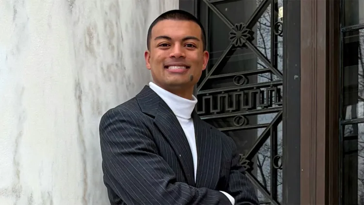 Young man in dark suit jacket and white shirt standing in front of marble wall smiling