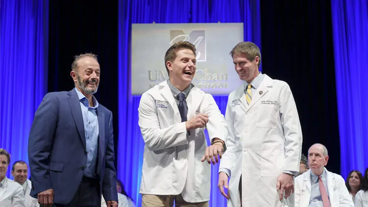 Three people on stage, two in white coats smiling and one in a suit, during a ceremony.
