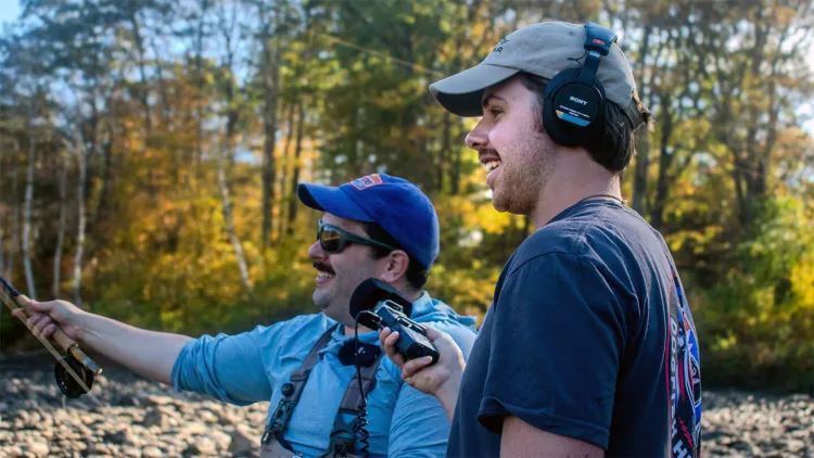 Man in blue cap and shirt fishing by river standing next to young man with recording equipment 