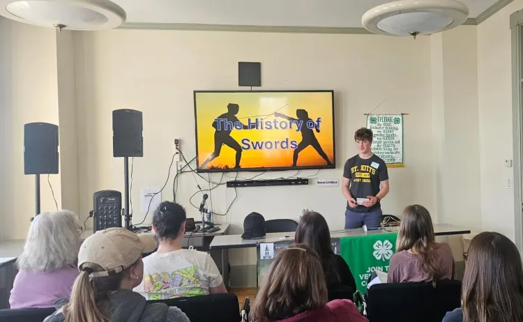 A young person stands in front of a seated crowd next to a screen with an orange and yellow backdrop, the silhouettes of two fencers and text reading the history of swords.
