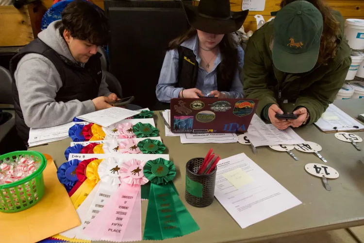 Three people at a table that has horse show ribbons, a laptop, peppermints on it