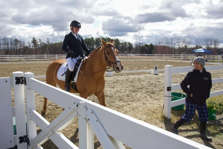 A person rides a horse through a white gate and a second person smiles up at the horse and rider