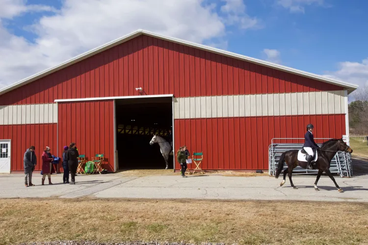 A red barn against a blue sky with a couple people on horses and many people on the ground.