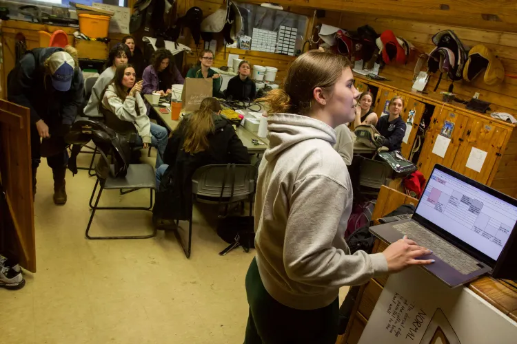 A young adult at a computer with a spreadsheet on it in front of a group of young adults in a tack room