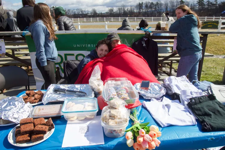 Two people sit snuggled up under a blanket in front of a table with cookies, brownies and flowers on it. A horse ring is in the background.