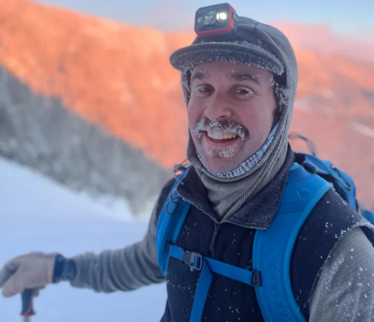 person with frost-covered facial hair and dressed in full winter gear stands on a mountainside in winter