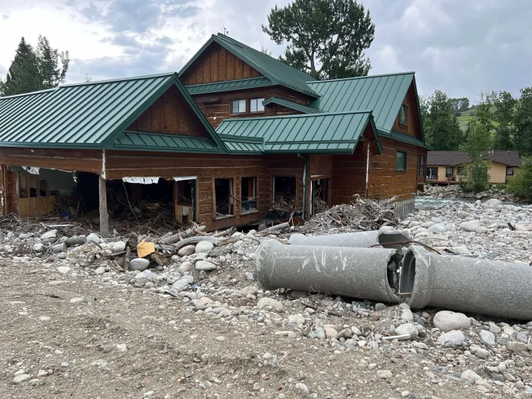 A heavily damaged wood-sided, green-roofed building with large rocks and a culvert on the ground.