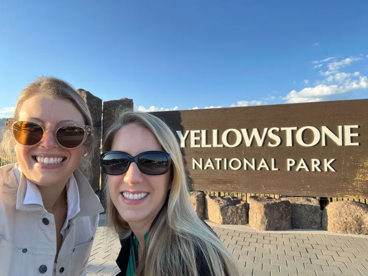 Two women in sunglasses smile by a brown sign reading yellowstone national park