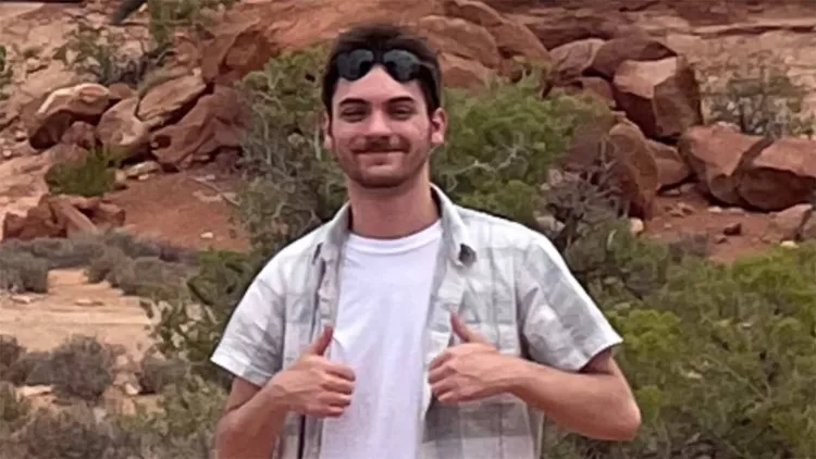 Young man with brown hair standing in front of rocky slope with greenery
