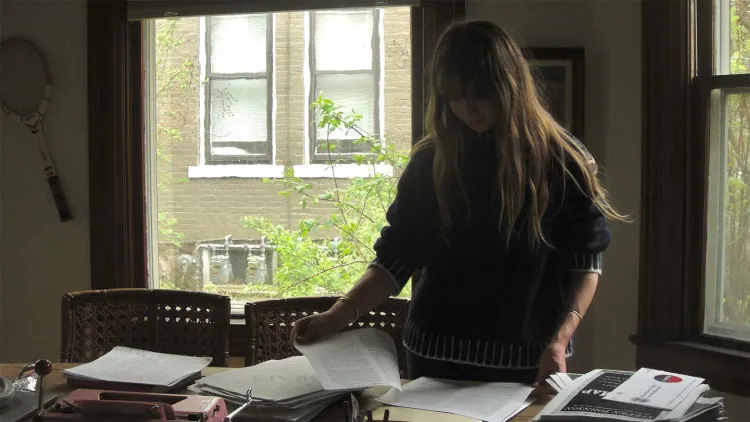 Young woman with long light hair standing in front of windows looking at papers on desk